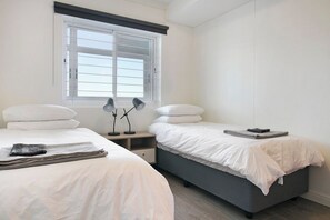 A hotel room with two beds, a window with blinds, a bedside table with a lamp, and a folded towel on the left bed.