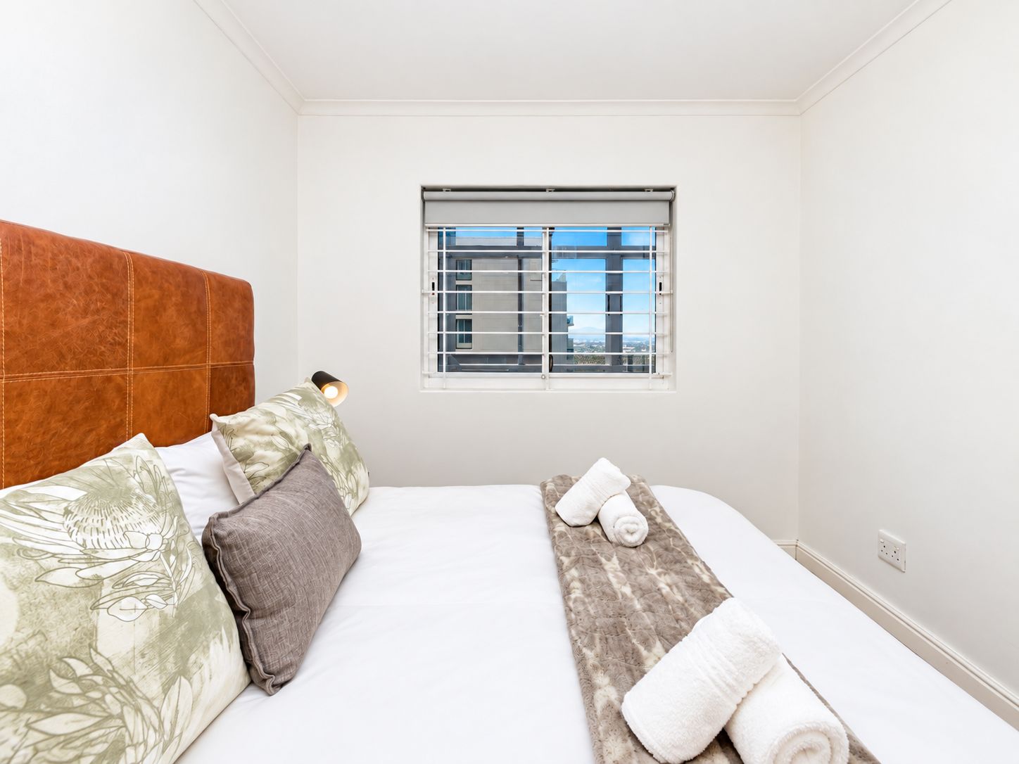 Un dormitorio con cama, almohadas, una lámpara de noche, una ventana con persianas y una toalla sobre la cama.