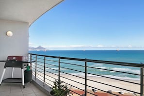 Een balkon met een barbecue, een bloempot en uitzicht op zee en strand.