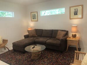 A living room with a brown sectional sofa, a glass coffee table, a patterned rug, a wooden side table with a lamp, and two framed pictures on the wall.
