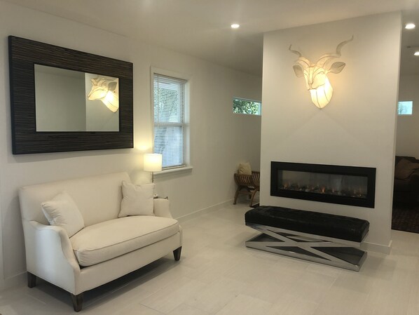 A modern living room with a white sofa, a fireplace, a wall-mounted mirror, and a decorative light fixture.