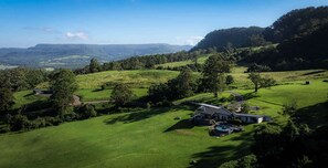 A large house with a swimming pool, surrounded by lush green fields and trees, with a view of distant hills.
