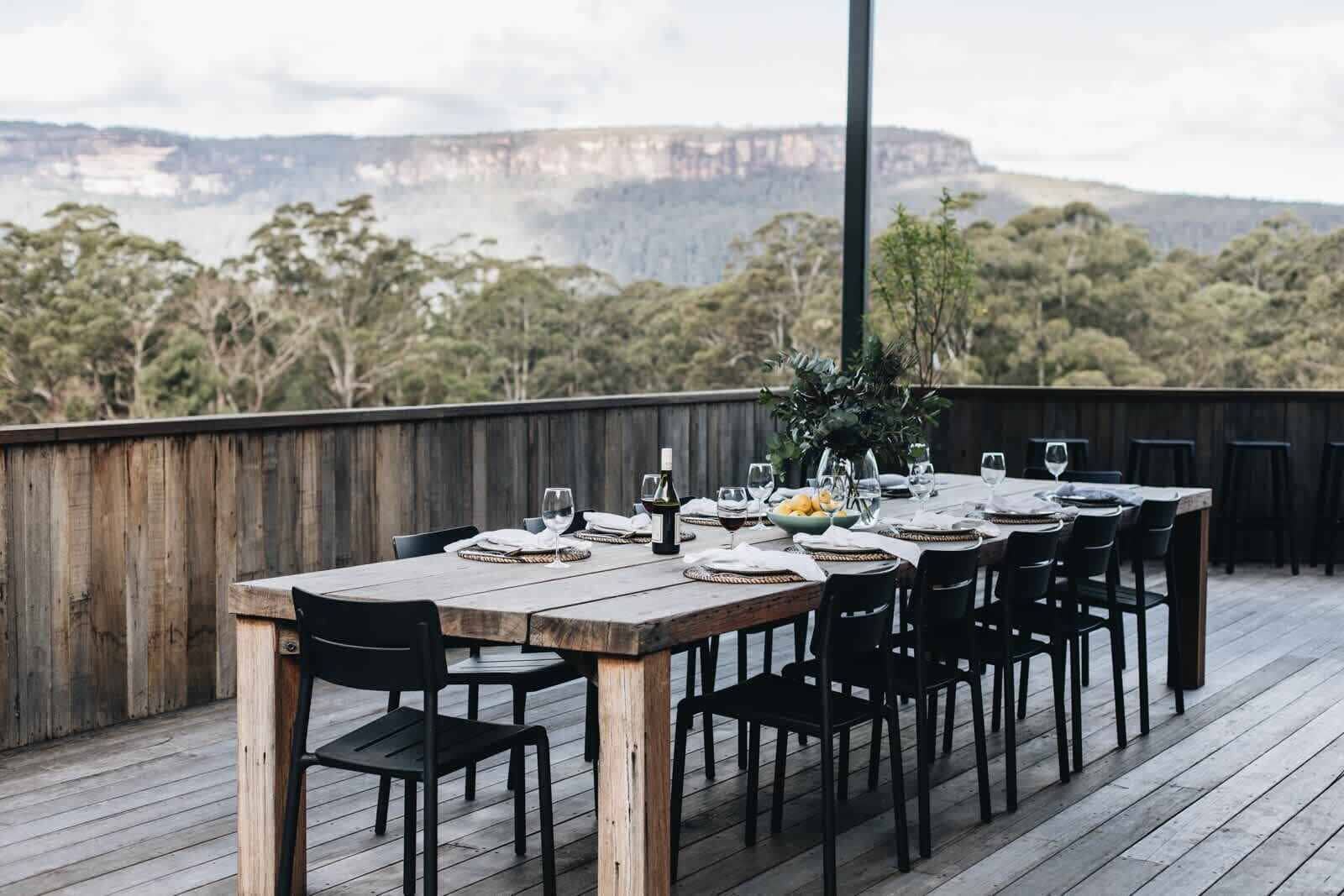 Una terraza de madera con una mesa lista para comer, con vista a un paisaje natural con árboles y un acantilado lejano.
