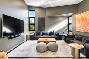 A modern living room with a flat-screen TV, grey sofas, a wooden coffee table, and a large window.