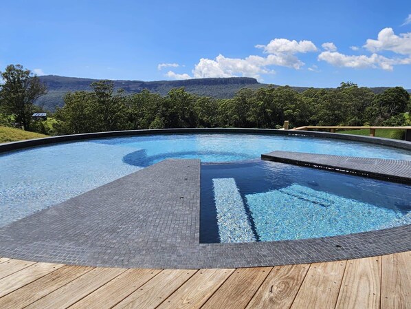 A swimming pool with a wooden deck, surrounded by trees and a view of hills.
