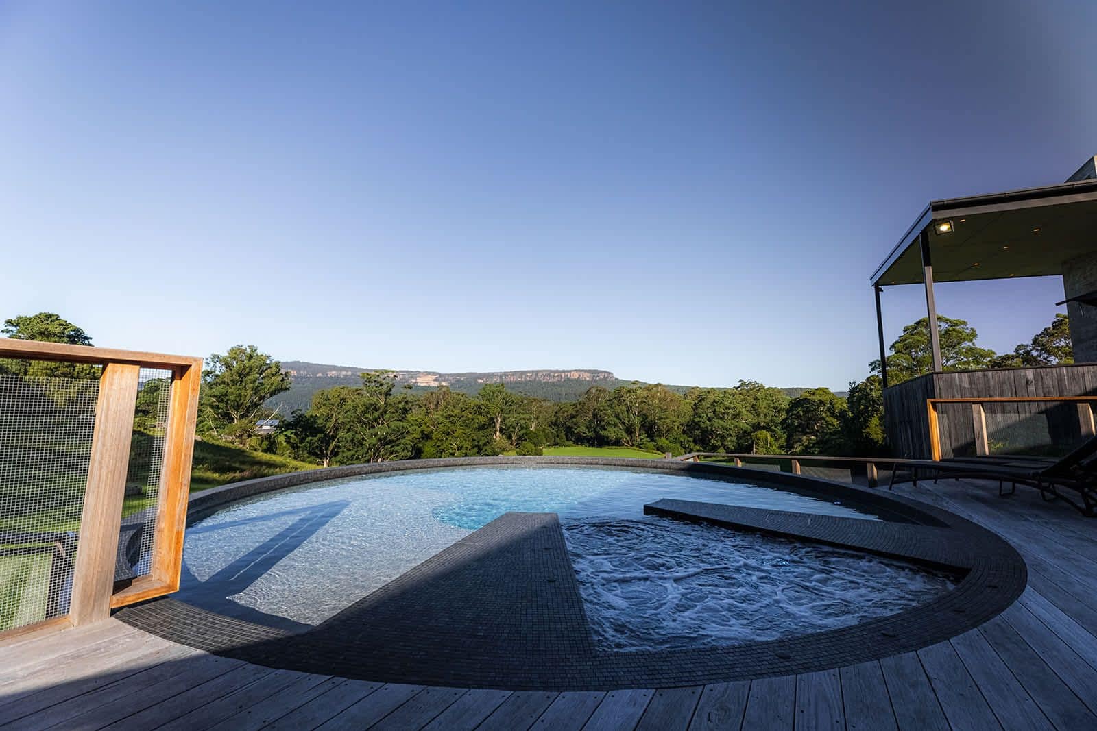 A swimming pool with a wooden deck, surrounded by greenery and a view of distant hills.