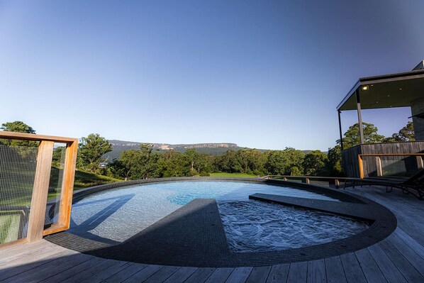 A swimming pool with a wooden deck, surrounded by greenery and a view of distant hills.