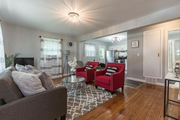 A modern living room with a grey sofa, red armchairs, and a coffee table. A kitchen is visible in the background.