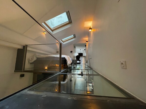 A narrow hallway with a glass floor leading to a bedroom with a bed and a skylight.