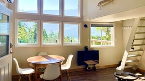 Una habitación con una mesa redonda de madera, sillas blancas, un televisor de pantalla plana y una escalera.