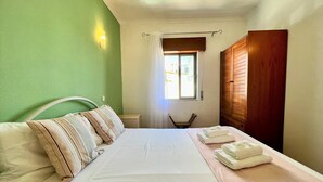 Una cama bien hecha con almohadas y toallas dobladas, una mesita de noche, una silla y una ventana con cortinas.
