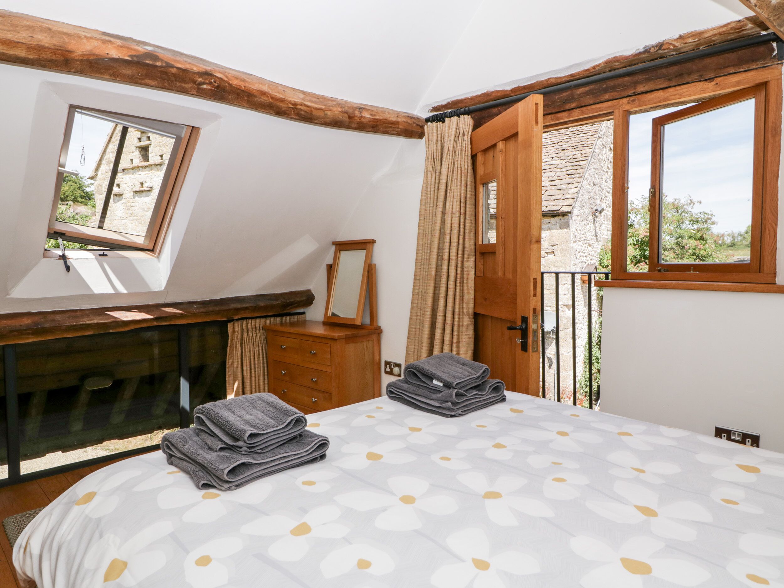 A bedroom with a sloped ceiling, a wooden dresser, a bed with a patterned bedspread, and a view of a stone building through a window.
