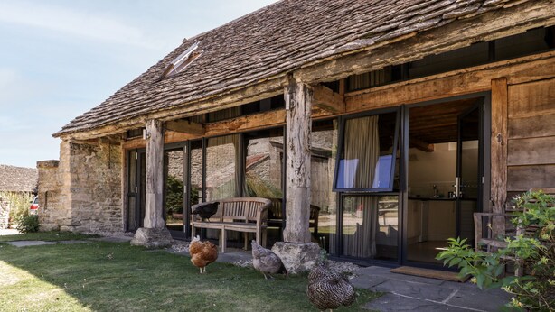 Een rustiek stenen gebouw met rieten dak, een houten bank en een glazen deur naar een moderne keuken.