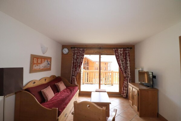Un salón acogedor con un sofá de madera, una mesita y un televisor. Se ve un balcón con barandas de madera a través de la ventana.