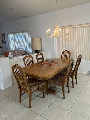 A dining area with a wooden table and six chairs, a chandelier, and a window with blinds.