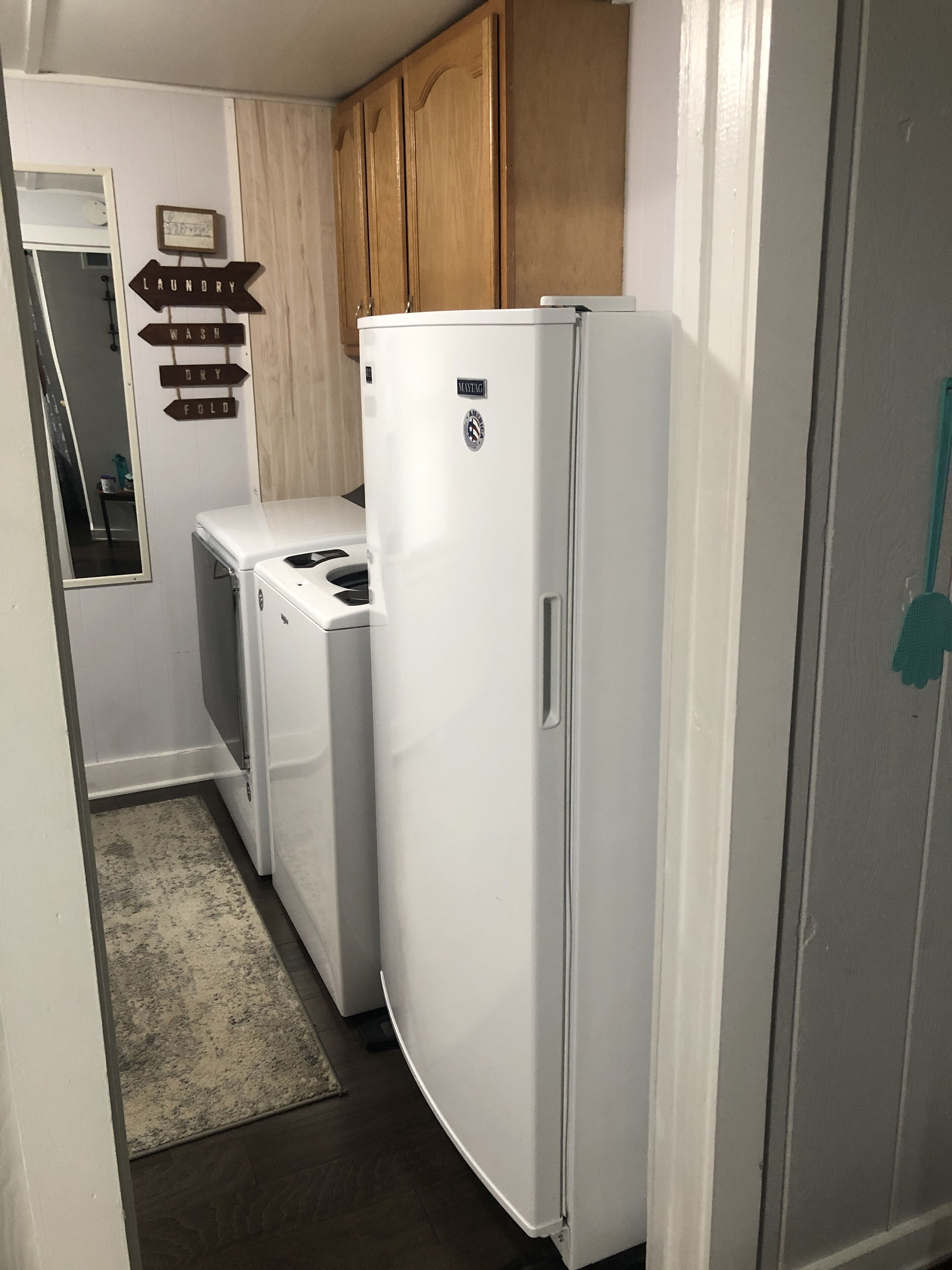 A compact kitchen with a white refrigerator, a washing machine, and a dryer. There is a wooden cabinet above the appliances and a small mirror on the wall.