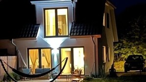 Una casa de dos pisos con luz interior visible, una hamaca y un coche estacionado.