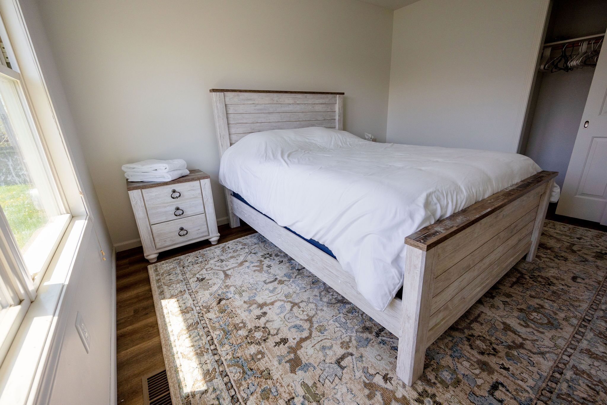 Un dormitorio con una cama de madera, una mesita de noche blanca con cajones, una alfombra estampada y una ventana.
