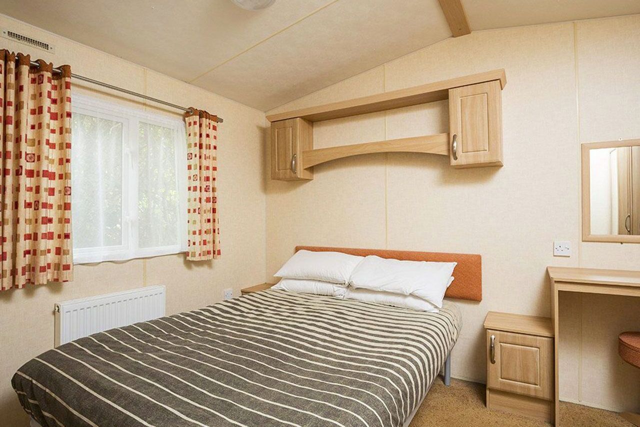 A bedroom with a bed, a window with curtains, a wooden cabinet, a bedside table, and a mirror.