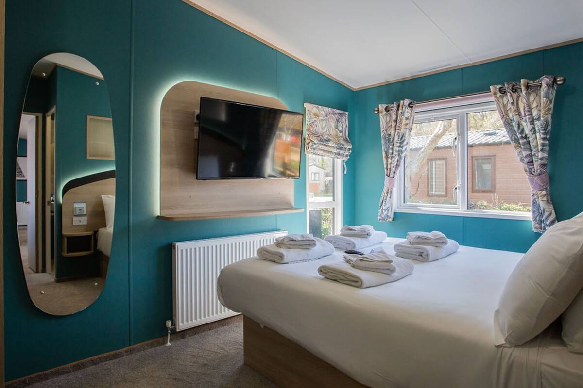 Une chambre avec des murs de couleur turquoise, un grand lit avec du linge de lit blanc, un téléviseur à écran plat fixé au-dessus du lit, un miroir et une fenêtre ornée de rideaux à motifs.