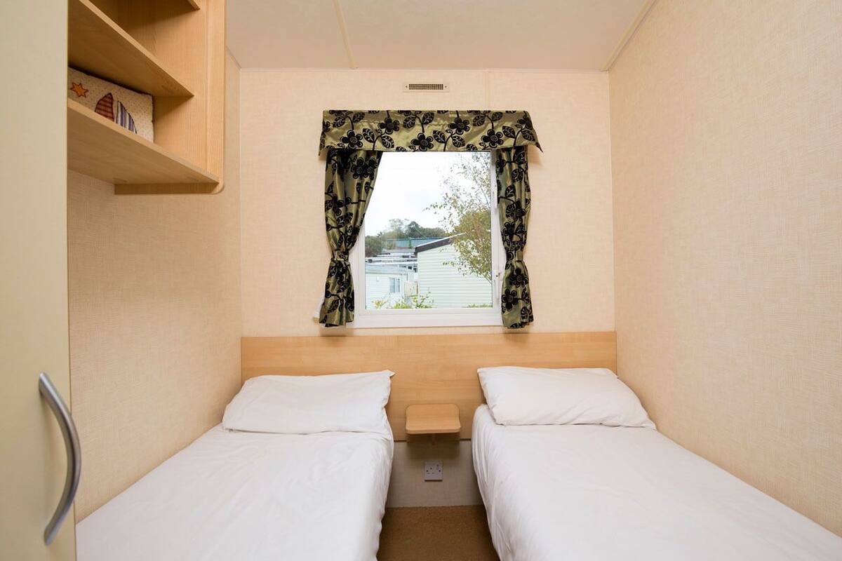 Une petite chambre d’hôtel composée d’une seule pièce, avec deux lits, une fenêtre ornée de rideaux à motifs floraux, une étagère en bois remplie de livres et une petite table placée entre les lits.
