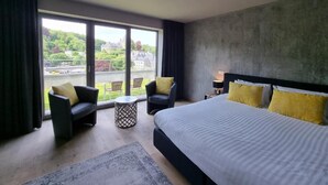 Habitación de hotel moderna con una cama grande, dos sillones, una mesita y un balcón con vistas a zonas verdes y edificios.