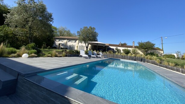 Une piscine avec des chaises longues, une maison avec un toit en tuiles et un ciel bleu azur.