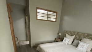 Une chambre à coucher avec un grand lit, une table de chevet, une petite table ornée d’un vase de fleurs et une fenêtre avec des stores.