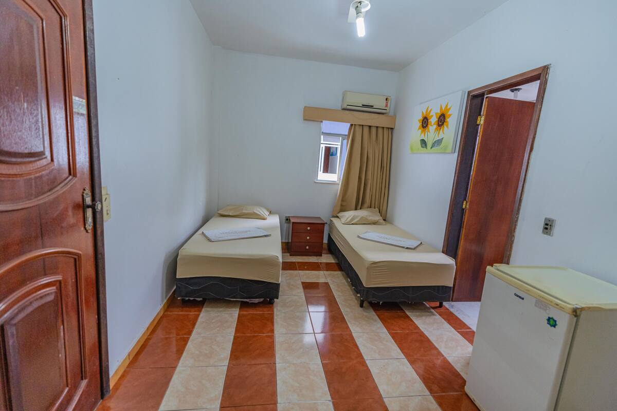 A small, clean room with two single beds, a wooden door, an air conditioning unit, a small window with curtains, and a refrigerator.