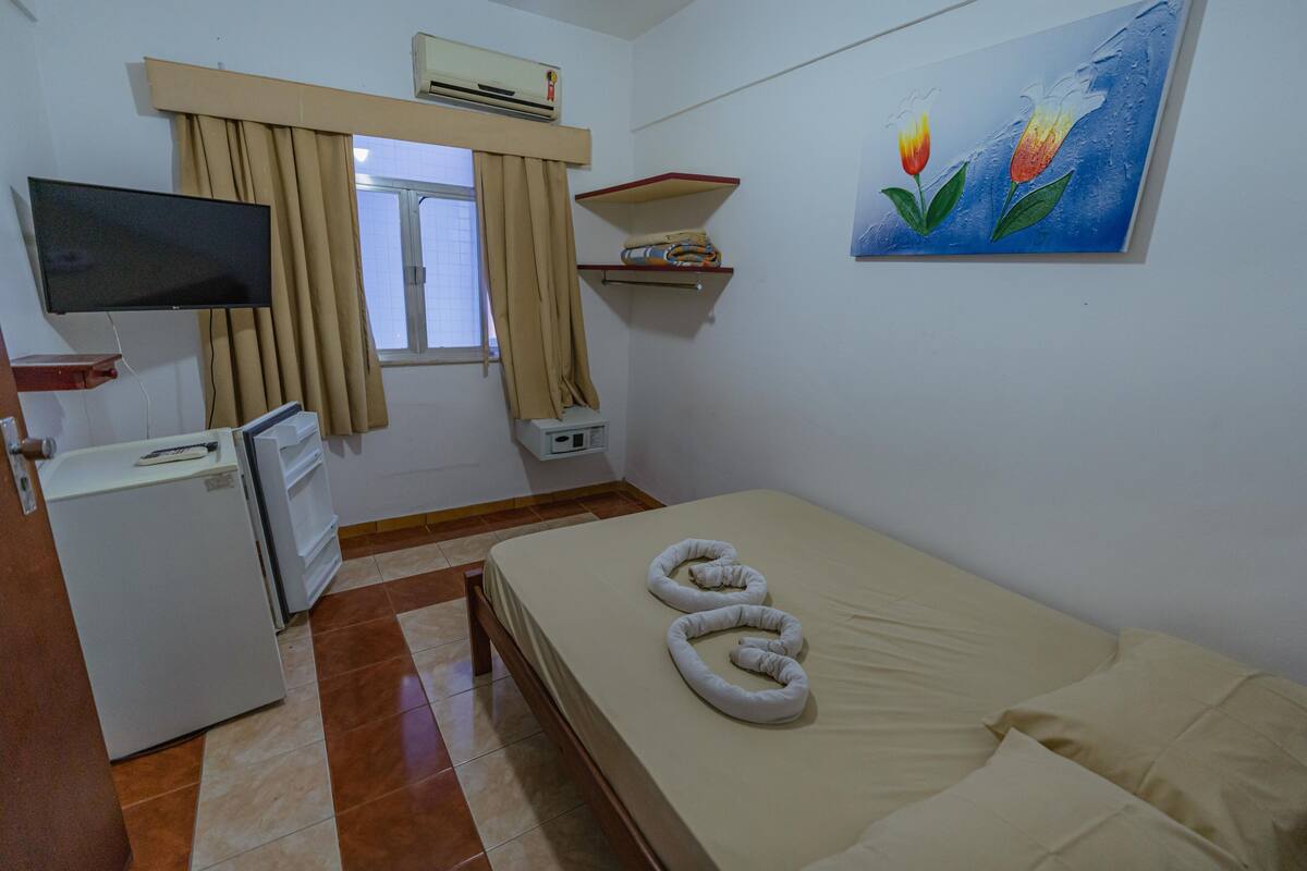 A room with a bed, a television, a refrigerator, a window with curtains, and a painting on the wall.