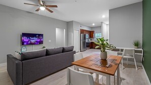 A modern living room with a sofa, dining table, and chairs, a TV mounted on the wall, and a ceiling fan. - Lovely newly built condo centrally located in TPA (Tampa)
