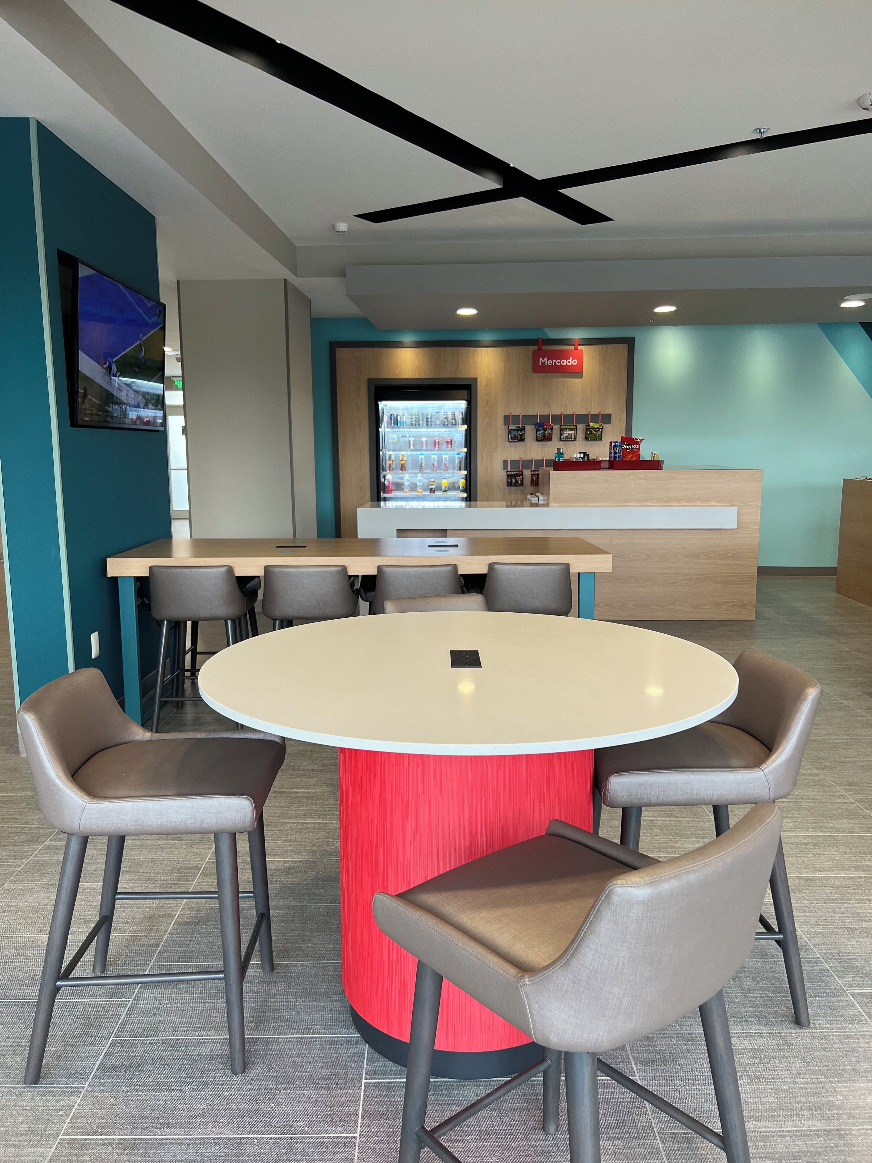 A modern indoor seating area with a round table and six chairs, a vending machine, and a counter with a 'Mercado' sign.