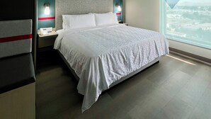 Una habitación de hotel moderna con una cama grande, dos mesitas de noche con lámparas, vistas a la ciudad a través de un ventanal amplio y un cabecero a rayas rojas.