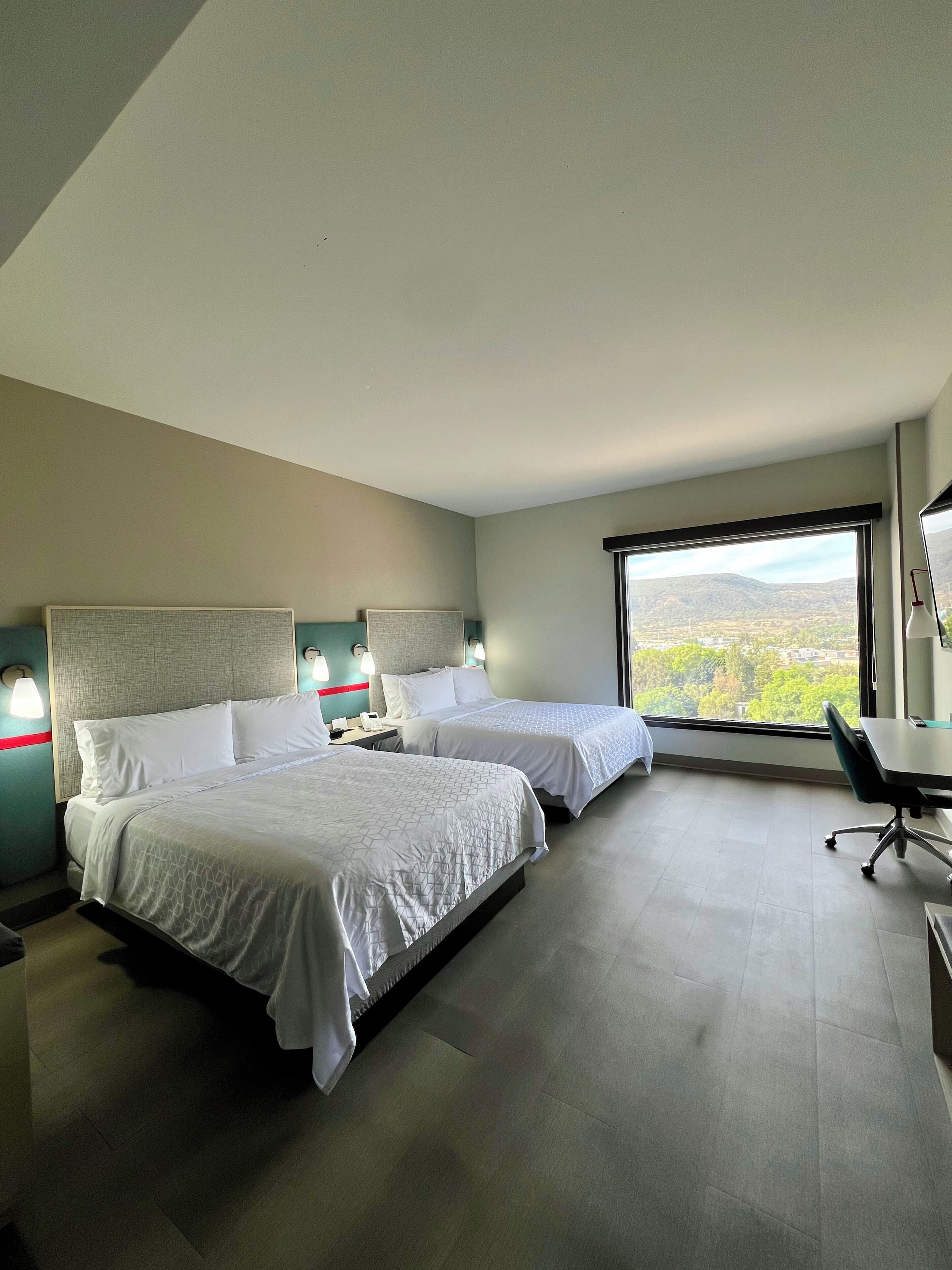Ein Hotelzimmer mit zwei Betten, einem Schreibtisch und einem großen Fenster mit Blick auf die Landschaft.