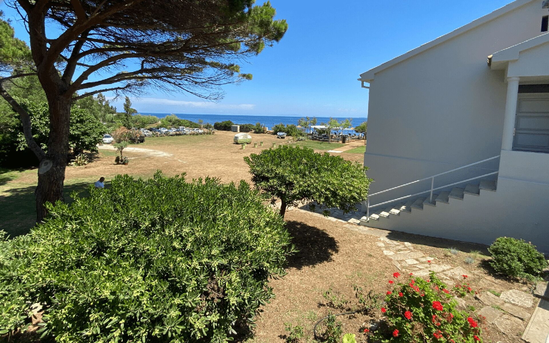Une propriété résidentielle avec vue sur la mer, un jardin agrémenté d’arbustes et d’un petit arbre, et un bâtiment blanc doté d’un escalier.