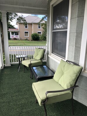 Varanda com duas cadeiras estofadas verdes e uma pequena mesa, uma janela grande e vista para uma casa na rua.
