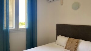 A hotel room with a bed, a window with blue curtains, an air conditioning unit, and a wall-mounted light. - Welcome to Domaine U Pozzu (SISCO)