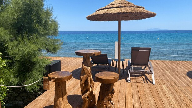 A wooden deck with a thatched umbrella, two round tables, and two chairs overlooking the sea. - Welcome to Domaine U Pozzu (SISCO)