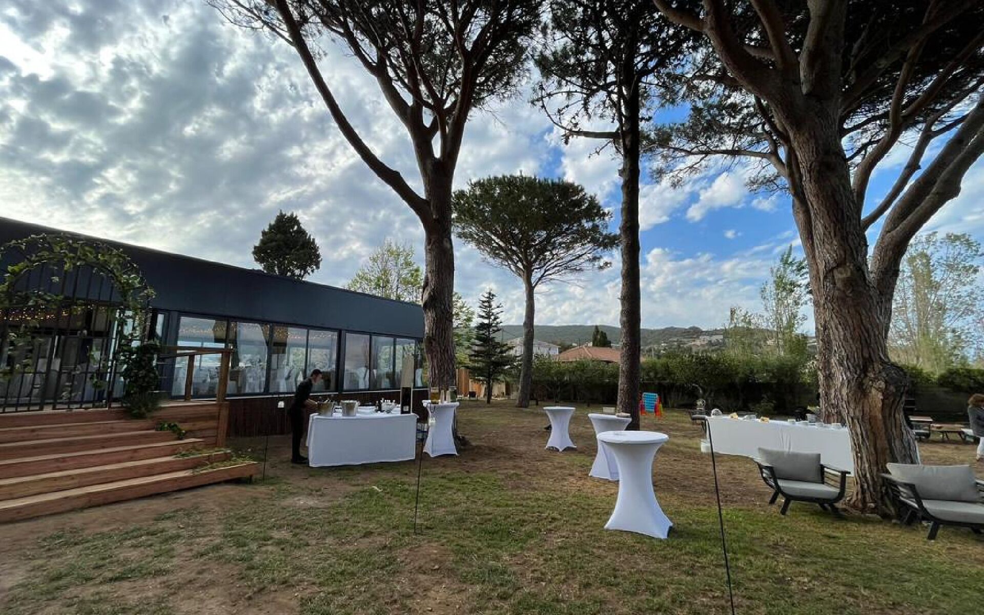 An outdoor event space with white round tables and chairs, a building with large windows, and a grassy area with trees.