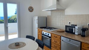 A compact kitchen with a gas stove, microwave, oven, and dishwasher. There is a dining table with chairs, and a view of the outdoors through a sliding door. - Welcome to Domaine U Pozzu (SISCO)