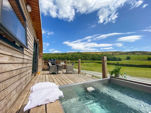 Une terrasse en bois avec un jacuzzi, un espace salon extérieur et une vue panoramique sur une vallée verdoyante.