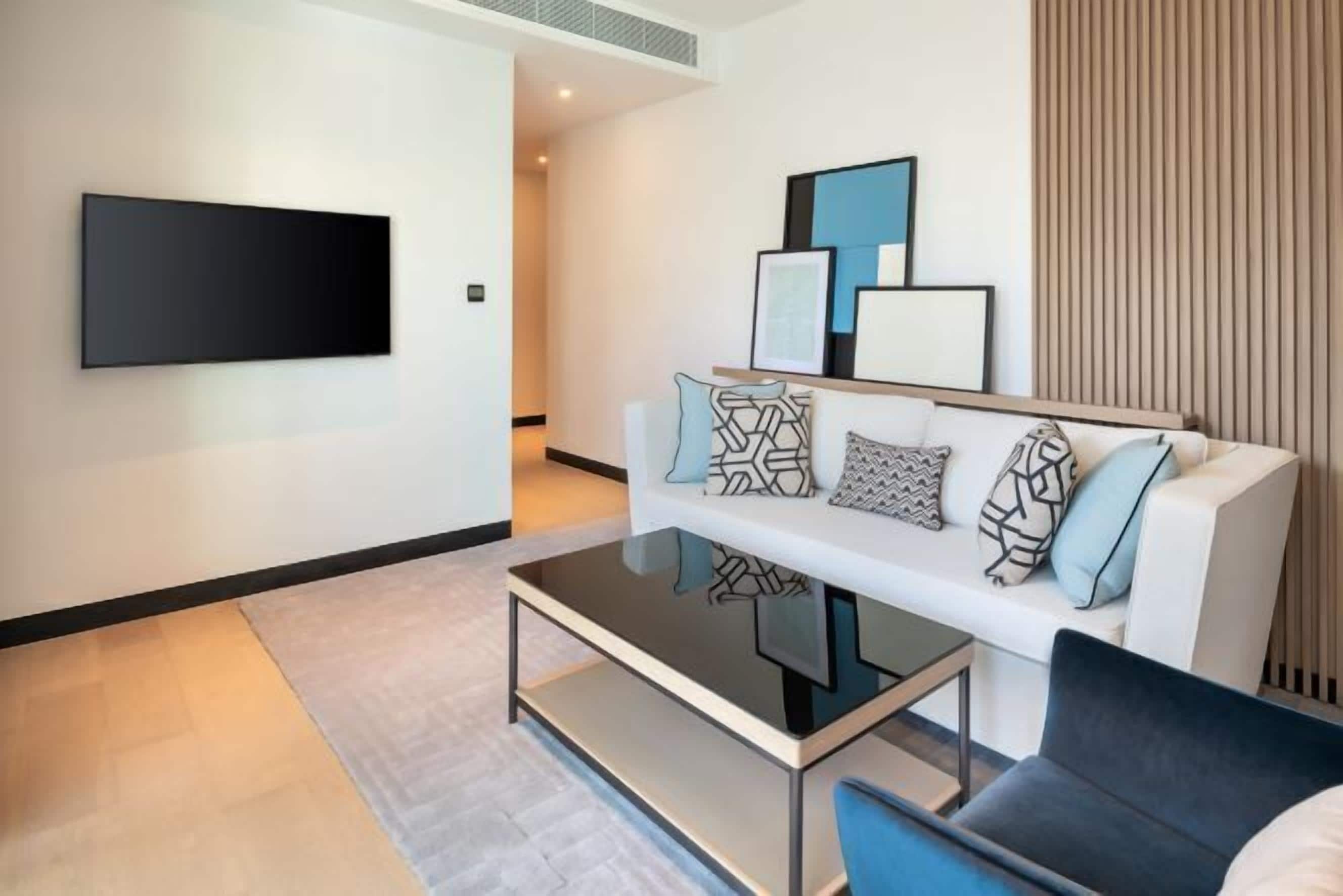 Una sala de estar moderna con una televisión de pantalla plana montada en la pared, un sofá blanco con cojines estampados, una mesa de centro de vidrio y un sillón oscuro.