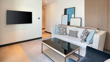A modern living room with a flat-screen TV mounted on the wall, a white sofa with patterned cushions, a glass coffee table, and a dark-colored armchair.