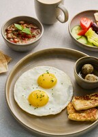 A plate with two sunny-side-up eggs garnished with chives, accompanied by a bowl of hummus, a small bowl of falafel balls, a plate of sliced cucumbers, tomatoes, and olives, and a piece of flatbread.