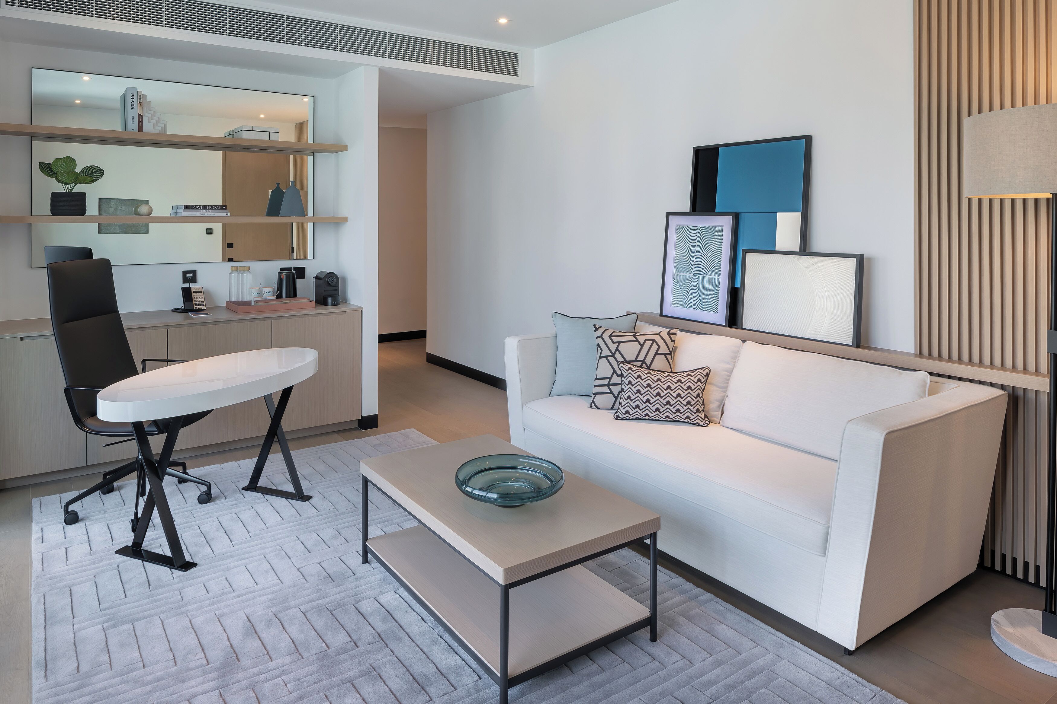 A modern living room with a white sofa, a coffee table, a dining table with chairs, and a kitchen area with shelves and a mirror.