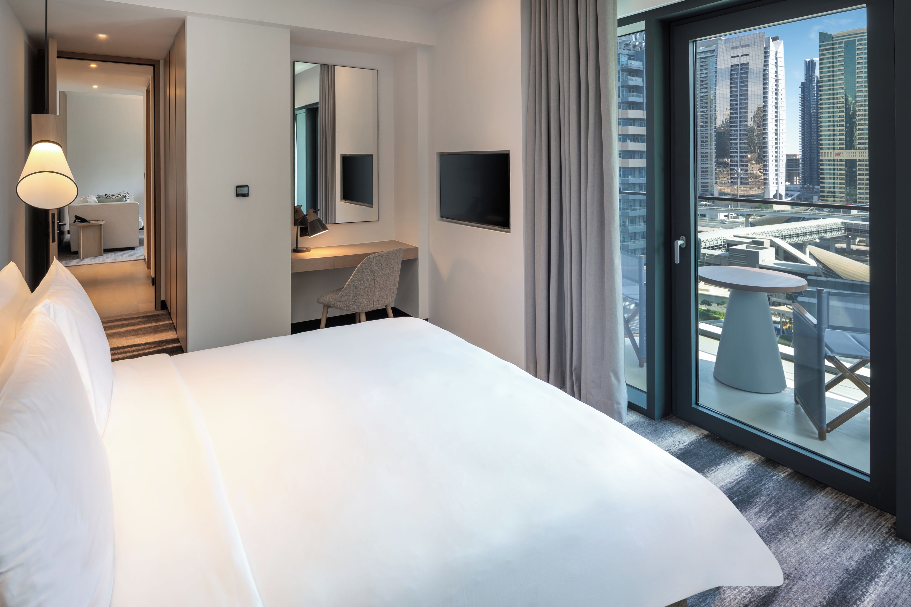 A hotel room with a large bed, a desk, a chair, a TV, and a balcony with a city view.