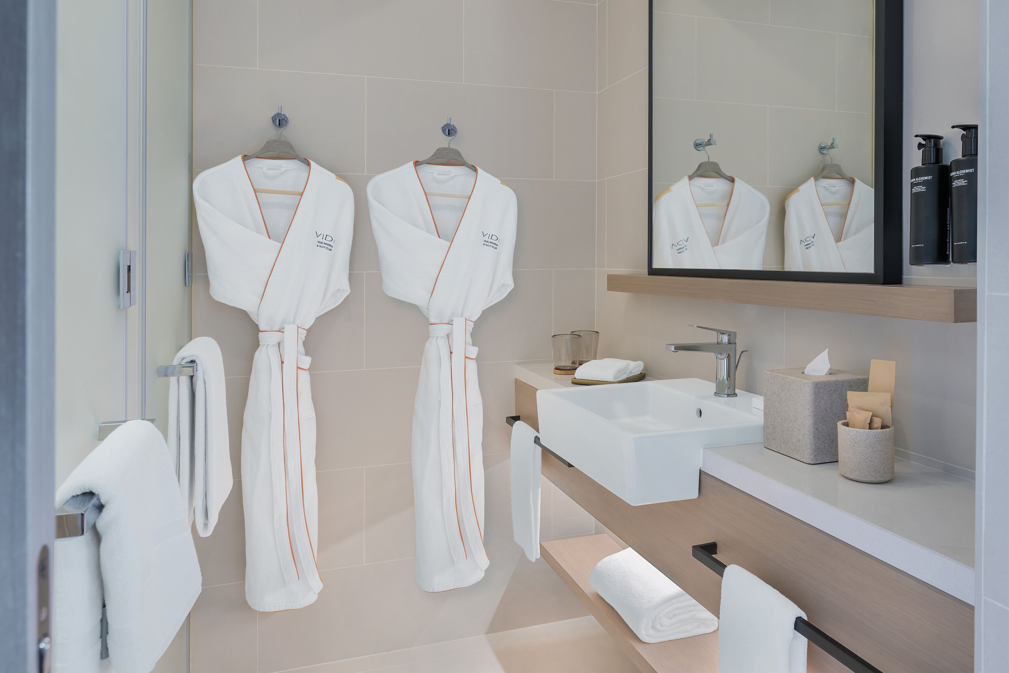 Ein modernes Badezimmer mit zwei weißen Bademänteln, die an Haken hängen, einem Waschbecken und einem Spiegel darüber.