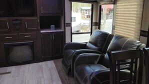 A compact living area with a dark wood cabinet, a flat-screen TV, a fireplace, and a leather sofa. - Modern Camper in the Smokies (Gatlinburg)