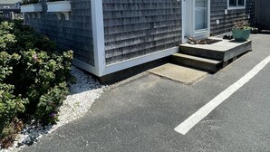 A small, single-story house with a grey shingle exterior and a white trim. It has a front door, a small porch, and a window.
