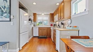 A narrow kitchen with wooden cabinets, a white refrigerator, and a dining area with a table and chairs.